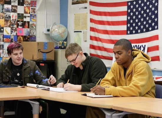 Three students sitting at a table in a classroom, with an American flag in the background. One student in a yellow hoodie is focused on writing, the middle student in glasses is also writing, and the third student with dyed hair is looking up.
